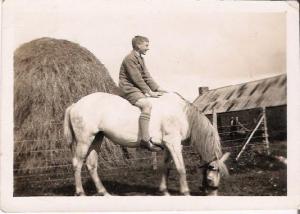 Black and white photograph of a white boy in school uniform riding a white pony bareback, smiling, with a hayrick and outhouse in the background.