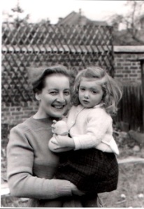 Black and white photograph of a smiling white woman holding her daughter, a blonde girl aged three, in her arms. They are standing in a garden and the girl is wearing a tartan skirt and holding a rubber duck.
