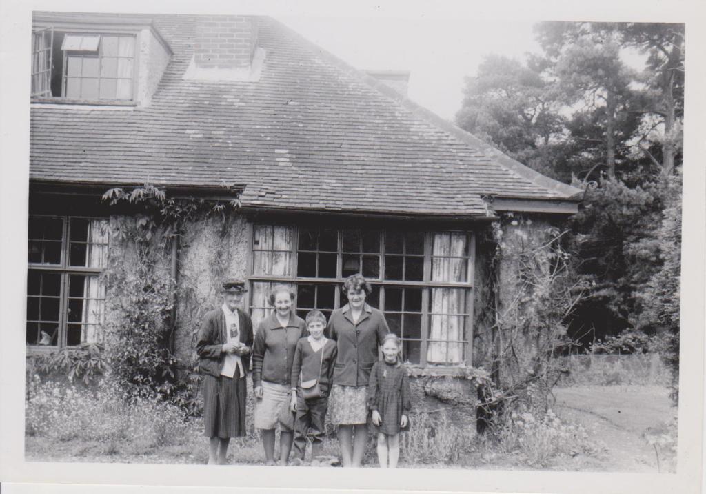 Black and white photograph taken in 1969 of a group of white people standing outside a 1920s house with trees behind it. There is an elderly woman in a hat, skirt, blouse and jacket; two women in their 40s  in skirts and cardigans and curled hair; a boy aged 11 in jeans and jumper with a shoulder bag, and a girl aged 8 in a tartan dress with fair hair in plaits. The house has square mullioned windows, a sloping tiled roof with a dormer window, and creepers climbing the walls.