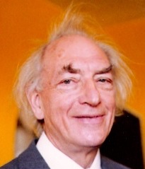 Colour photograph of an elderly white man smiling broadly, with a bald pate, messy grey hair and bushy eyebrows.