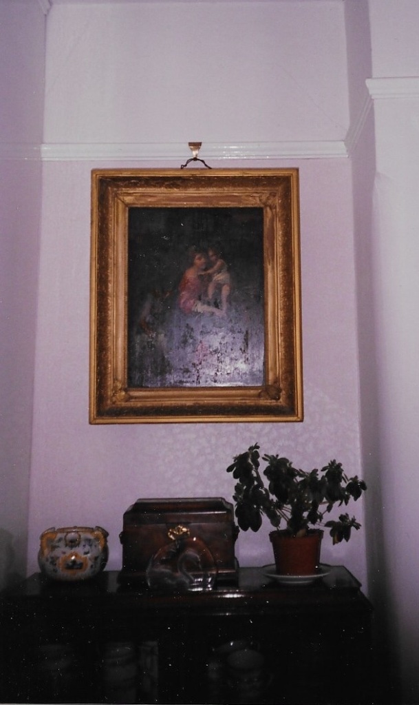 Photograph of an oil painting of a Madonna and child in an ornate gilt frame, hanging on a wall above a cabinet with ornaments and plants on top.
