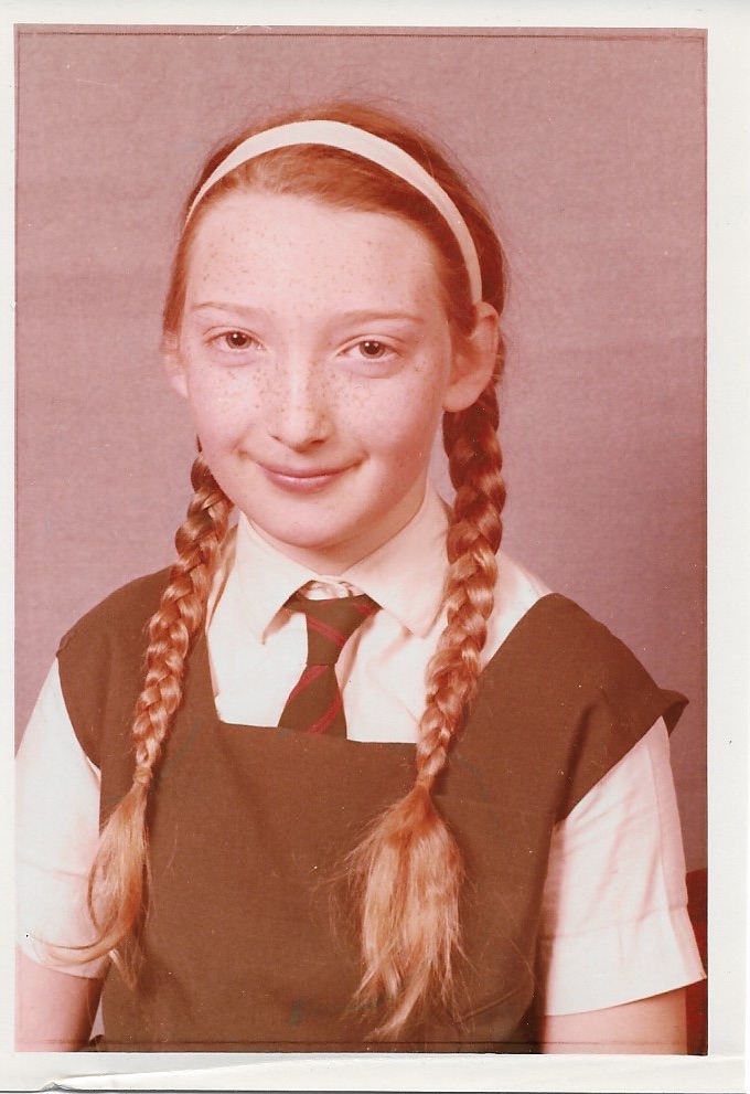 Colour photograph taken in 1972 of a white girl with brown hair in plaits with a white headband, a green school tunic, green and red striped tie  and white short sleeved blouse. 
