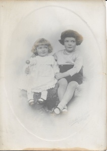 Black and white 1924 studio photograph of two smiling white children: a four year old boy with bushy dark hair wearing shorts, with his arm around a girl of about two with blonde hair wearing a white dress and holding a lollipop..