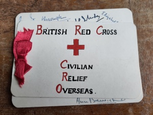 Hand made white greeting card with a red ribbon and "British Red Cross Civilian Relief Overseas" written in red and black, with a red cross. 