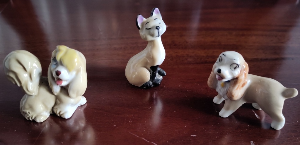 Three small china ornaments depicting characters from the Disney animated film The Lady and the Tramp: two dogs Peggy and Lady and a Siamese cat. 