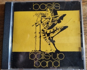 A Basil's Ballsup Band CD cover with a black and white caricature of a long haired man playing the flute, standing on one leg wearing a striped jumpsuit, on a yellow background. 