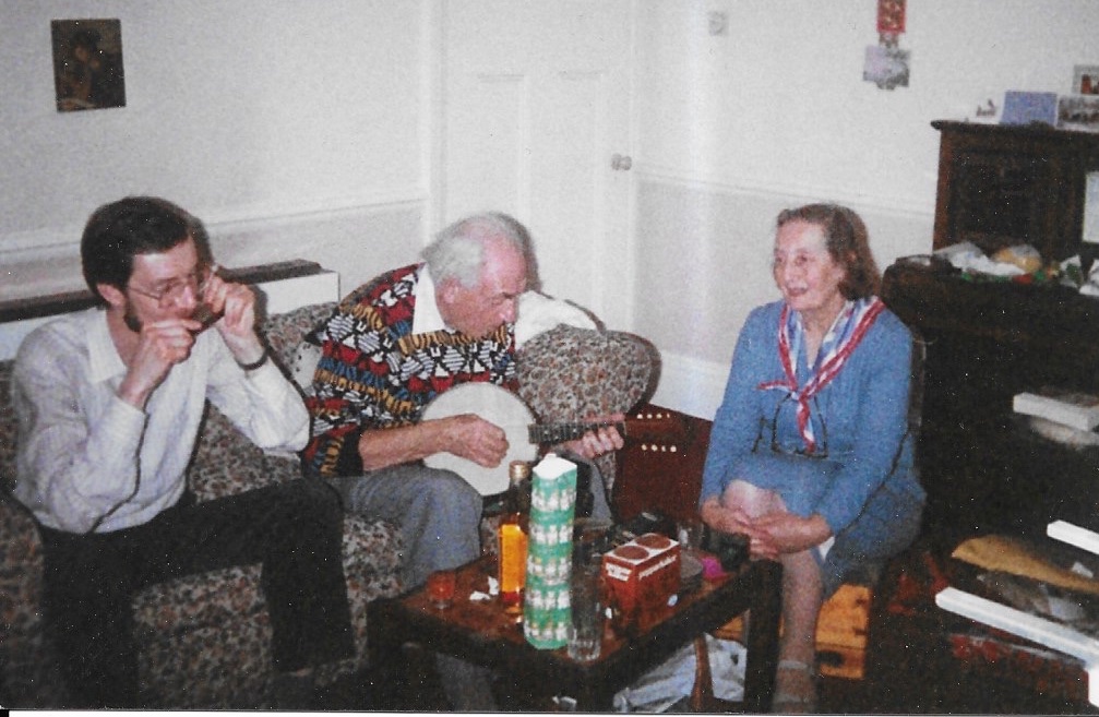 Three white people in a sitting room with Christmas decorations. On the sofa, a young man with dark hair and beard playing a harmonica, and an elderly man with white hair wearing a  colourful jumper playing a banjo-mandolin. A middle aged woman with brown hair in a blue dress sits in a chair listening to the music. There is an antique upright piano in the background.