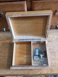 Wooden Music box with lid open, showing an empty compartment on the left side, and the rotating clockwork mechanism on the right side beneath a clear perspex lid. 